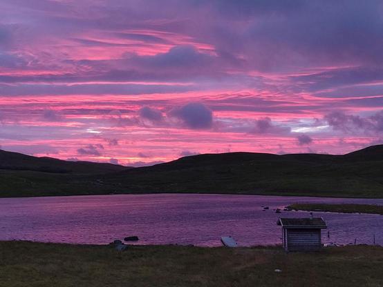 über einem See mit Hügeln dahinter leuchten nach Sonnenuntergang Wolkenstreifen in Pink und Lila