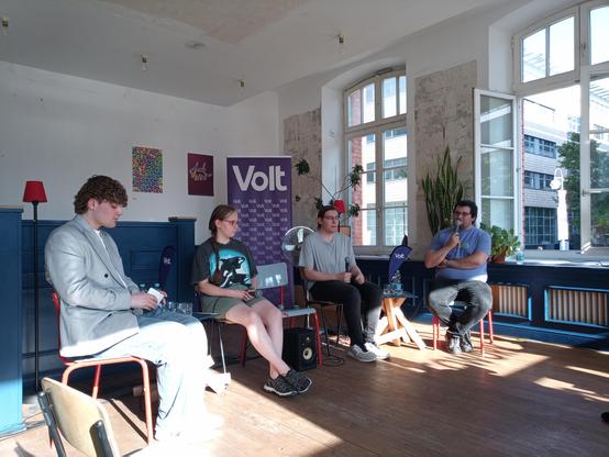 Auf der Bühne unsere 4 Gesprächsteilnehmer*innen zum ersten Thema Bildung.
Im Hintergrund ein Aufsteller mit Volt-Logo. Rechts im Bild ein großes geöffnetes Fenster.