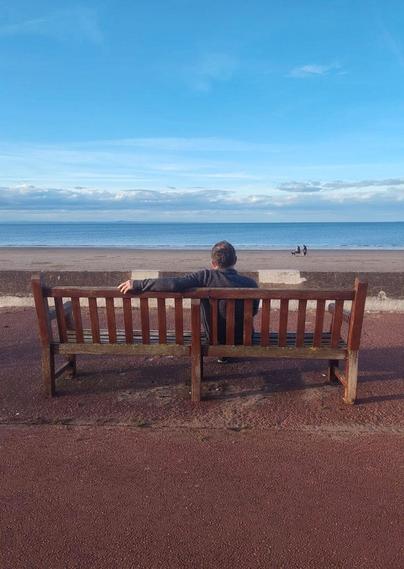 Foto von mir (von hinten) auf einer Bank am Strand, wie ich aufs Meer schaue.
